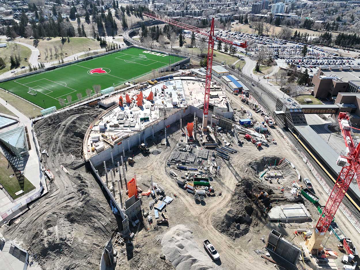 aerial view of the taylor family campus centre construction site, two large red cranes are surrounded on the ground with heavy vehicles and construction supplies, and large mounds of earth, a building foundation is partially constructed, the SAIT Trojans soccer field is in the background along with a large parking lot full of cars
