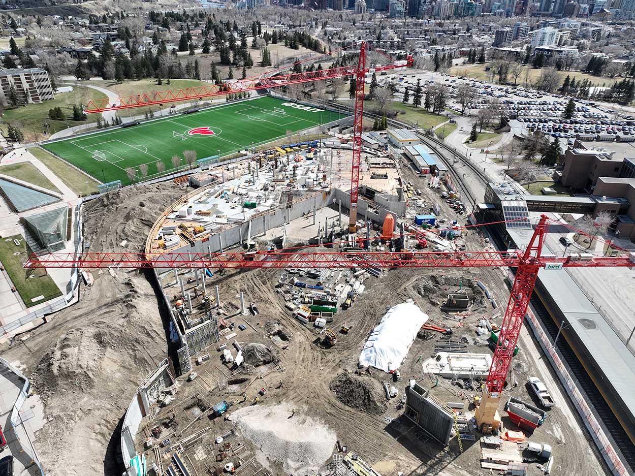 aerial view of the taylor family campus centre construction site, two large red cranes are surrounded on the ground with heavy vehicles and construction supplies, and large mounds of earth, a building foundation is partially constructed, the SAIT Trojans soccer field is in the background along with a large parking lot full of cars