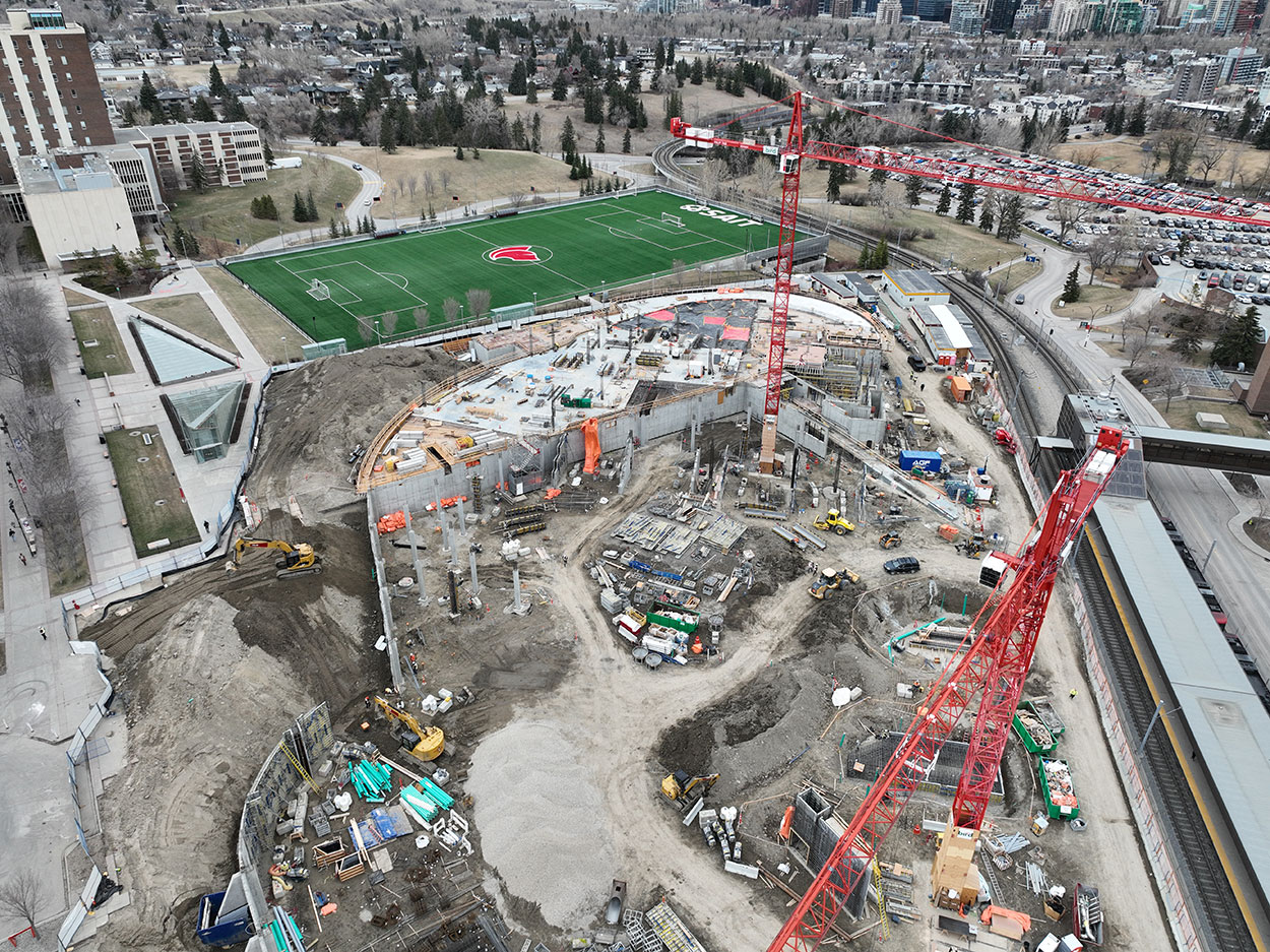aerial view of the taylor family campus centre construction site, two large red cranes are surrounded on the ground with heavy vehicles and construction supplies, and large mounds of earth, a building foundation is partially constructed, the SAIT Trojans soccer field is in the background along with a large parking lot full of cars