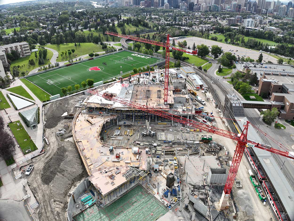 aerial view of the taylor family campus centre construction site, two large red cranes are surrounded on the ground with heavy vehicles and construction supplies, a building foundation and superstructure is partially constructed, the SAIT Trojans soccer field is in the background along with a large parking lot