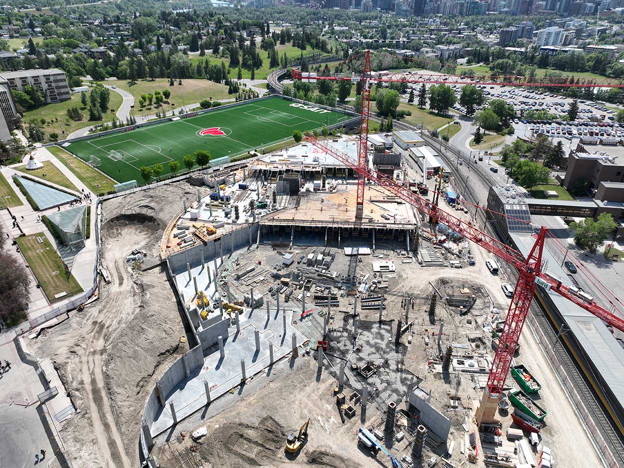 aerial view of the taylor family campus centre construction site, two large red cranes are surrounded on the ground with heavy vehicles and construction supplies, and large mounds of earth, a building foundation is partially constructed, the SAIT Trojans soccer field is in the background along with a large parking lot full of cars