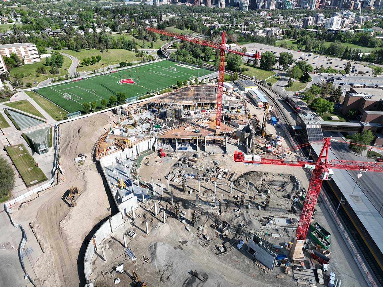 aerial view of the taylor family campus centre construction site, two large red cranes are surrounded on the ground with heavy vehicles and construction supplies, and large mounds of earth, a building foundation is partially constructed, the SAIT Trojans soccer field is in the background along with a large parking lot full of cars
