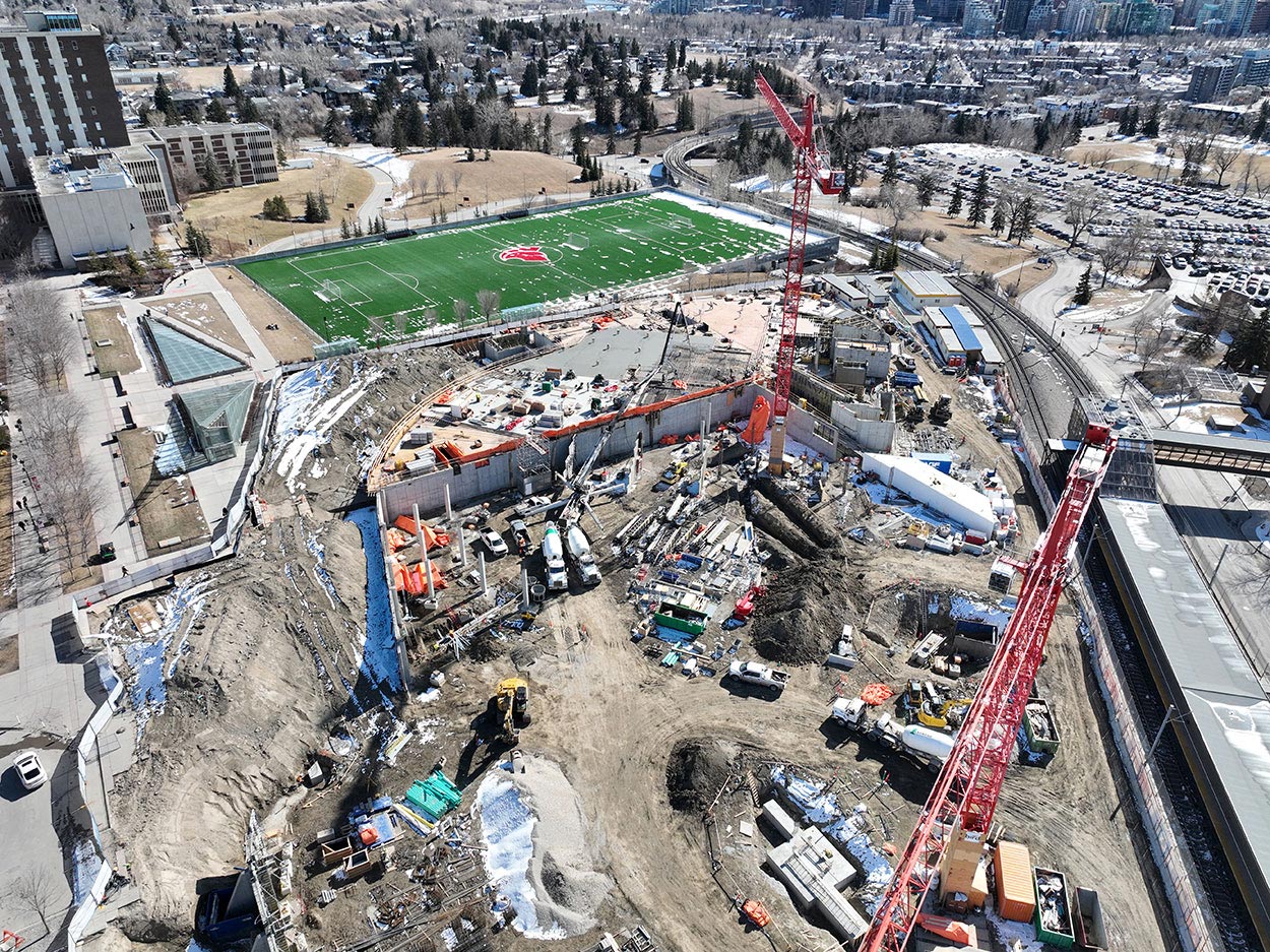 aerial view of the taylor family campus centre construction site, two large red cranes are surrounded on the ground with heavy vehicles and construction supplies, and large mounds of earth, a building foundation is partially constructed, the SAIT Trojans soccer field is in the background along with a large parking lot full of cars