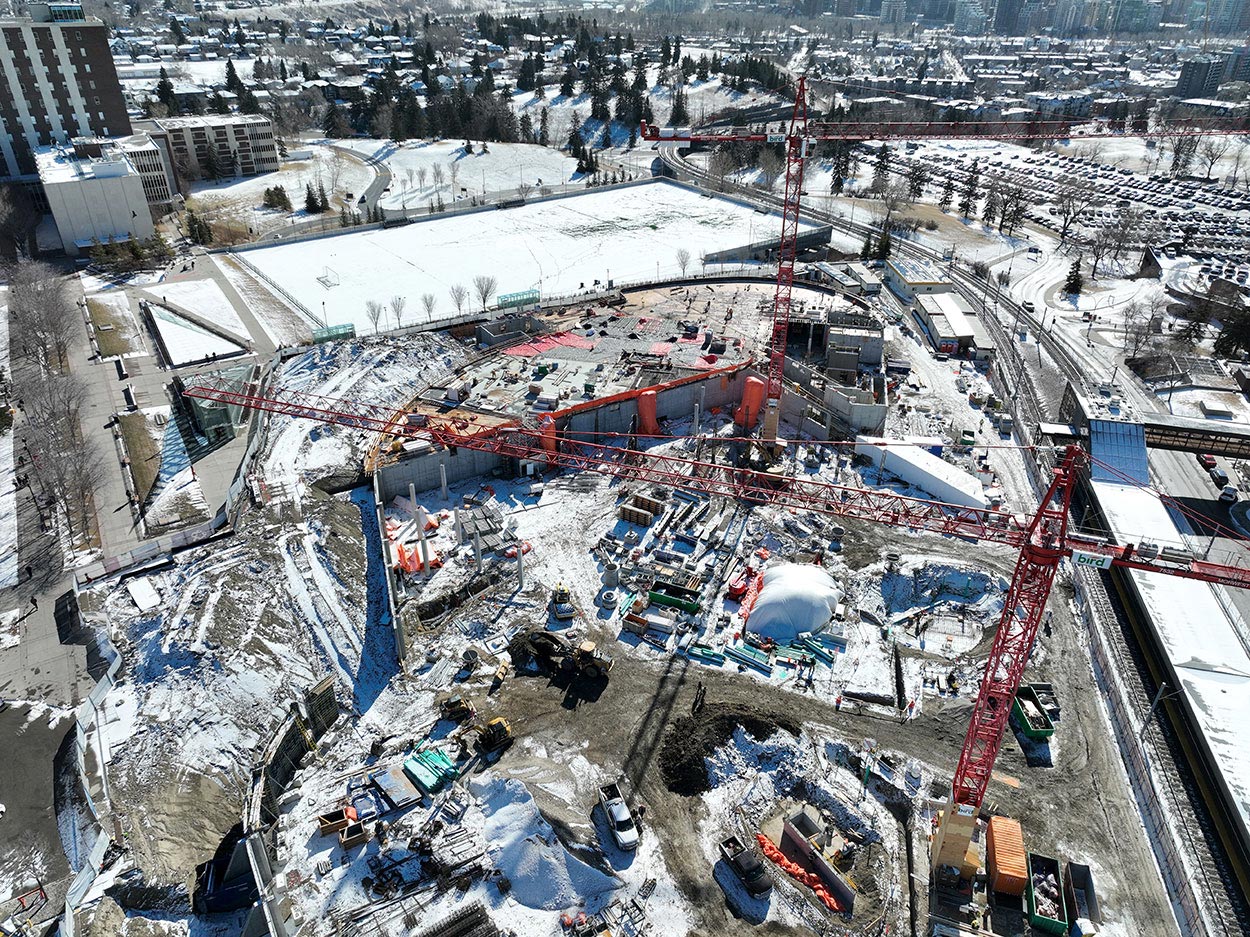 aerial view of the taylor family campus centre construction site, two large red cranes are surrounded on the ground, which is covered in snow, with heavy vehicles and construction supplies, and large mounds of earth, a building foundation is partially constructed, the SAIT Trojans soccer field is in the background, also covered in snow, along with a large parking lot full of cars
