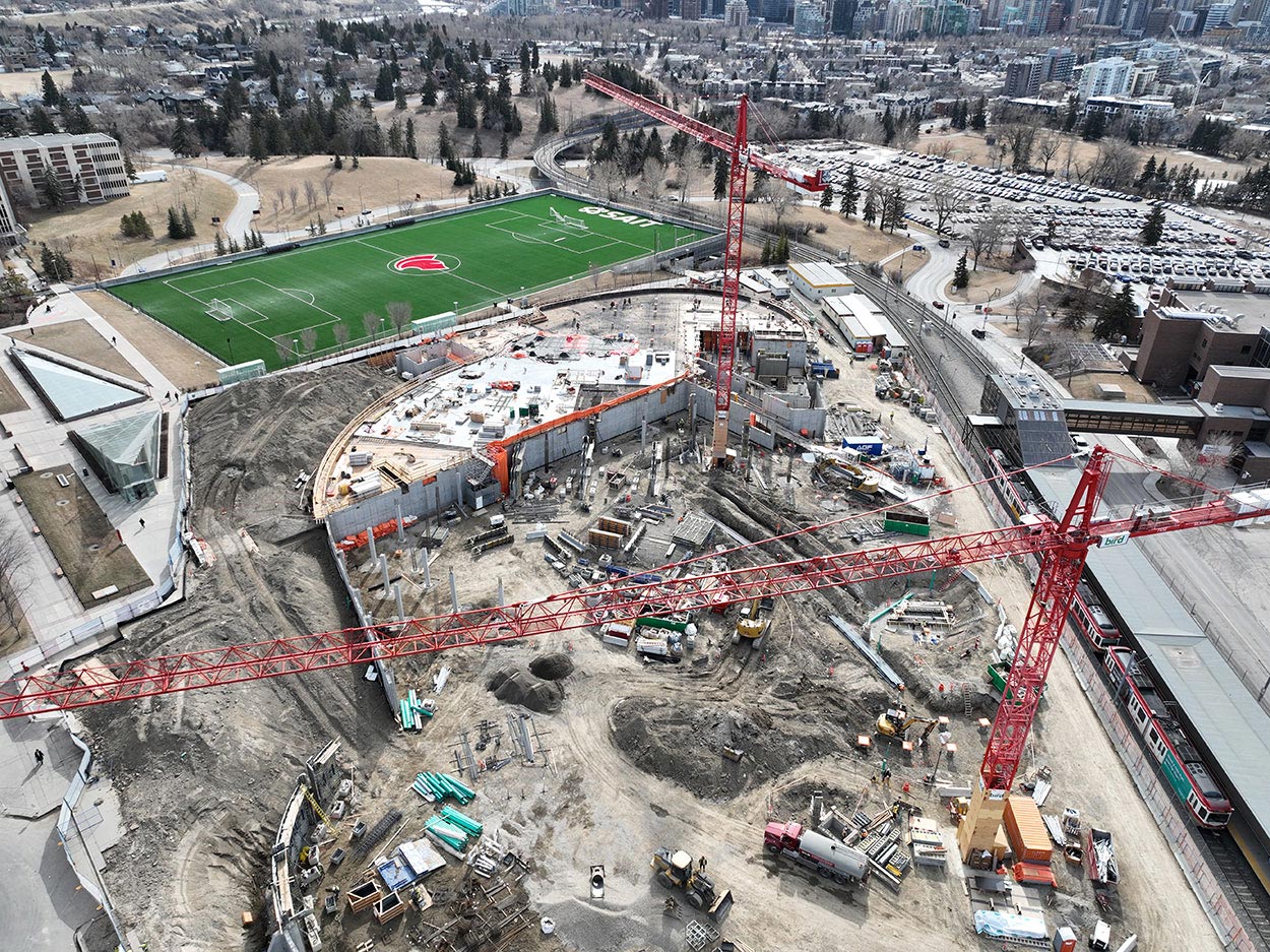 aerial view of the taylor family campus centre construction site, two large red cranes are surrounded on the ground with heavy vehicles and construction supplies, and large mounds of earth, a building foundation is partially constructed, the SAIT Trojans soccer field is in the background along with a large parking lot full of cars