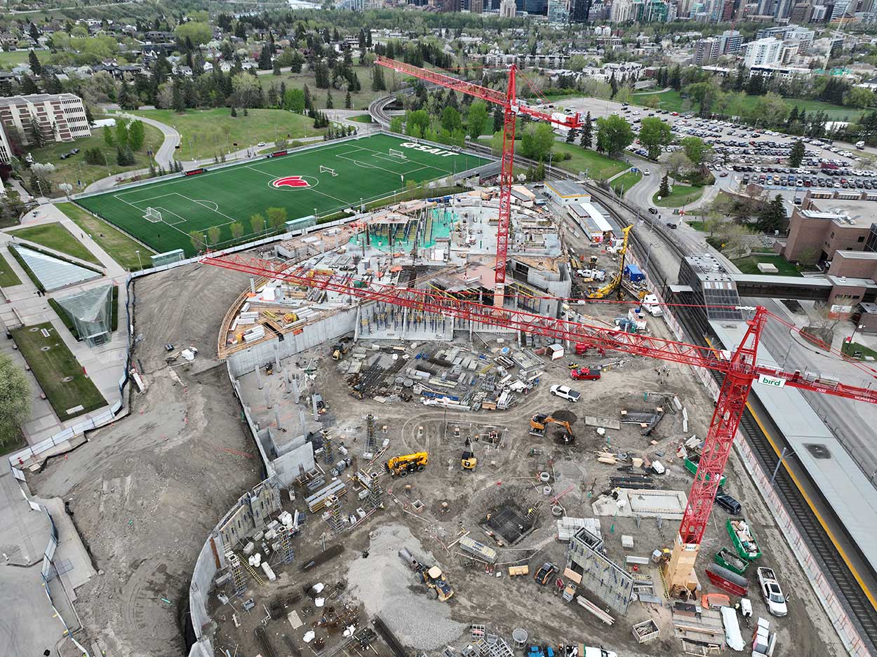 aerial view of the taylor family campus centre construction site, two large red cranes are surrounded on the ground with heavy vehicles and construction supplies, and large mounds of earth, a building foundation is partially constructed, the SAIT Trojans soccer field is in the background along with a large parking lot full of cars