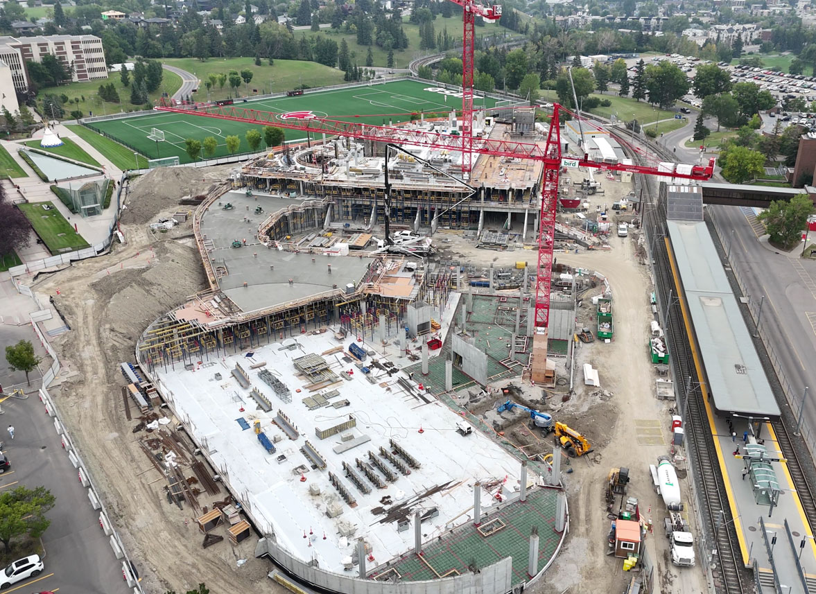 aerial view of the taylor family campus centre construction site, two large red cranes are surrounded on the ground with heavy vehicles and construction supplies, a building foundation and superstructure is partially constructed, the CTrain station and SAIT Trojans soccer field are in the background