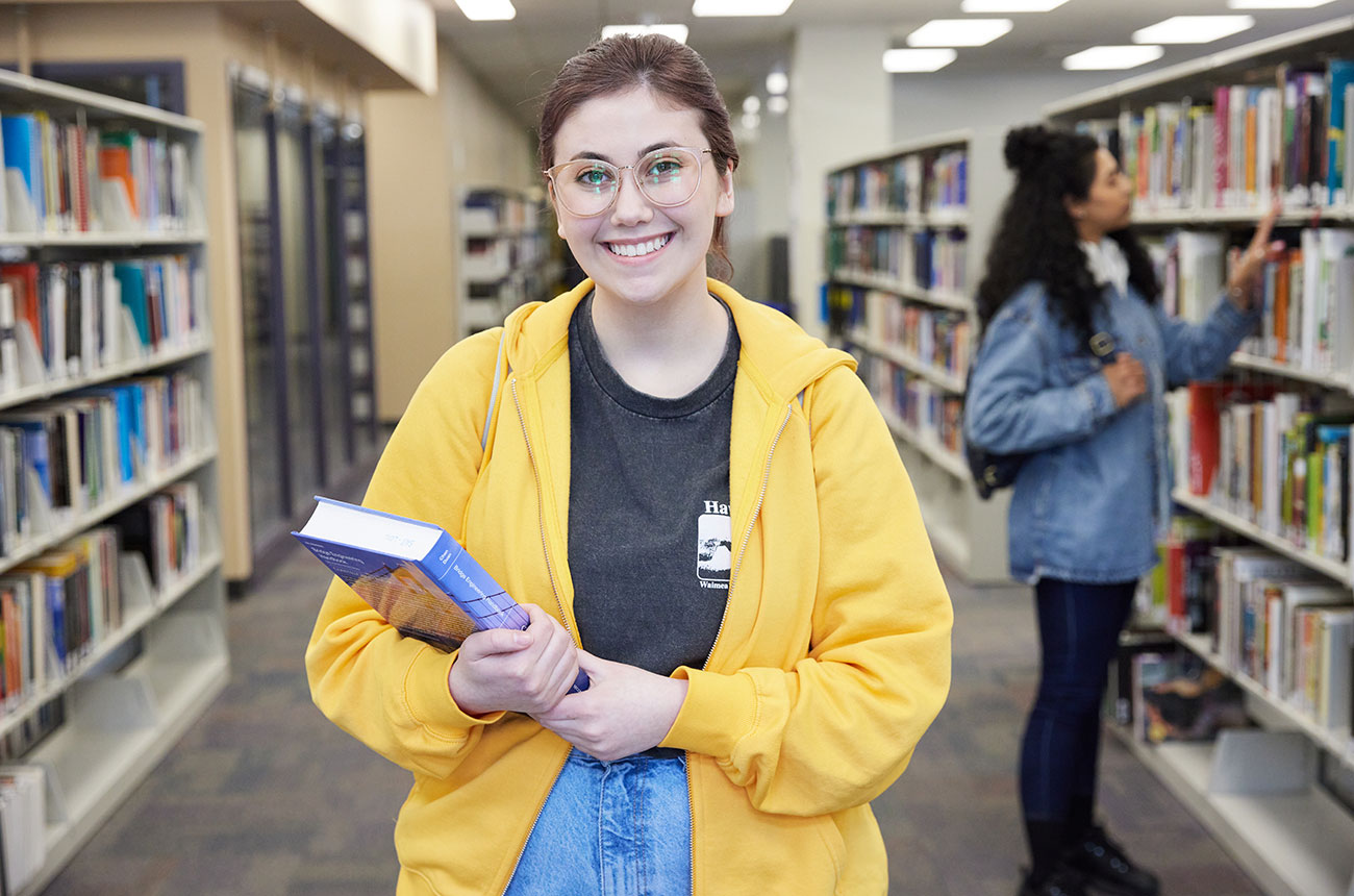 A woman wearing a bright yellow hoodie smiles while holding a textbook inside the Reg Erhardt Library.