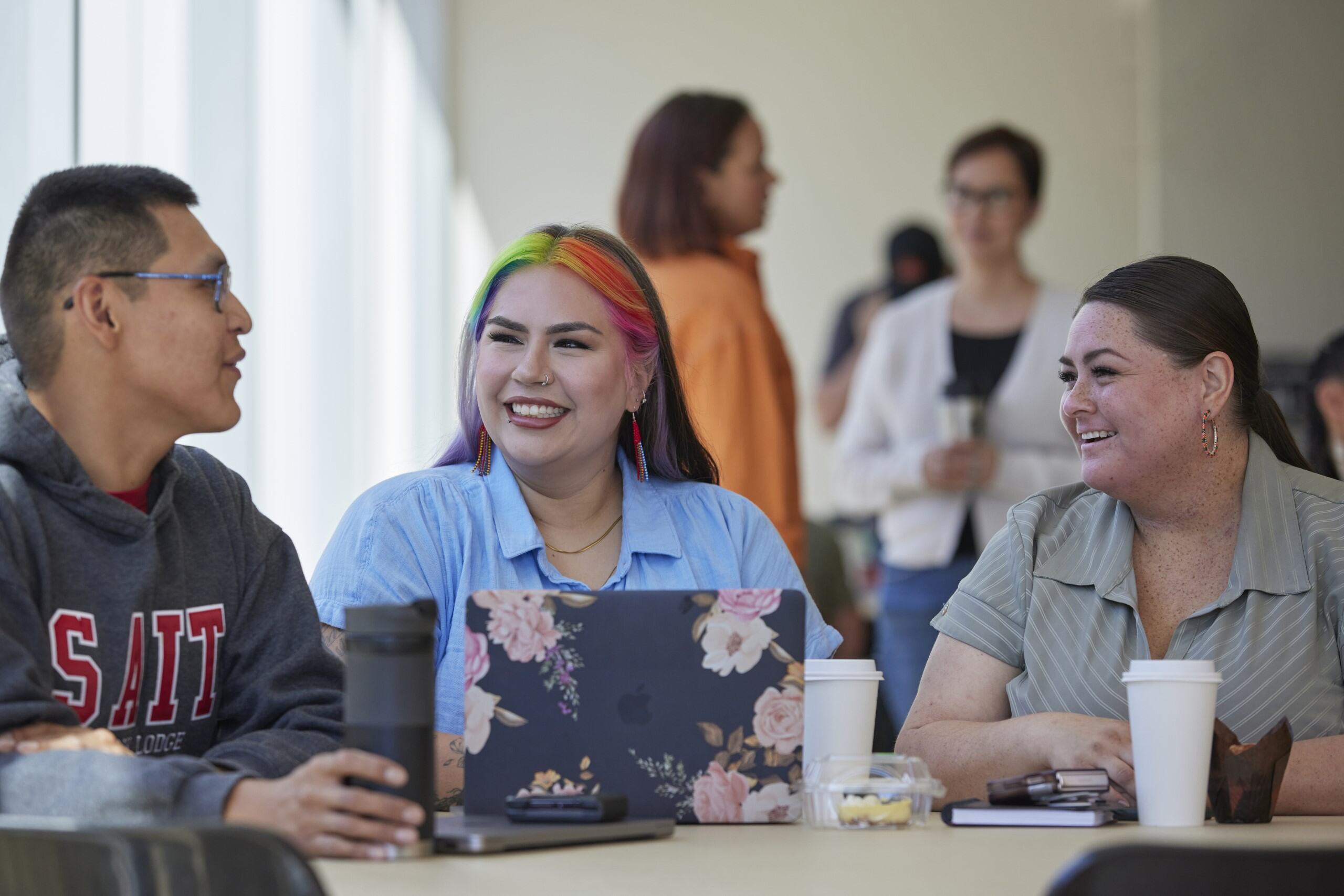 An Indigenous woman wearing colourful earrings smiles at others off-camera. She's sitting in Chinook Lodge Resource Centre.