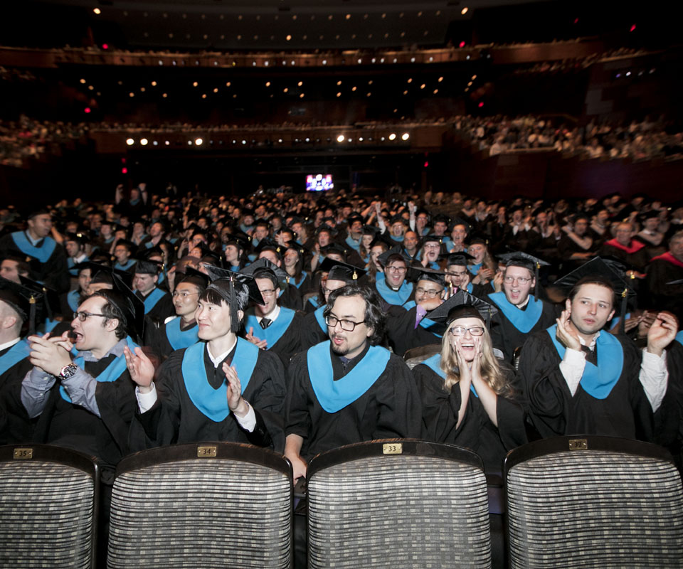 A large group of graduates in caps and gowns with turquoise hoods, celebrating at a graduation ceremony.