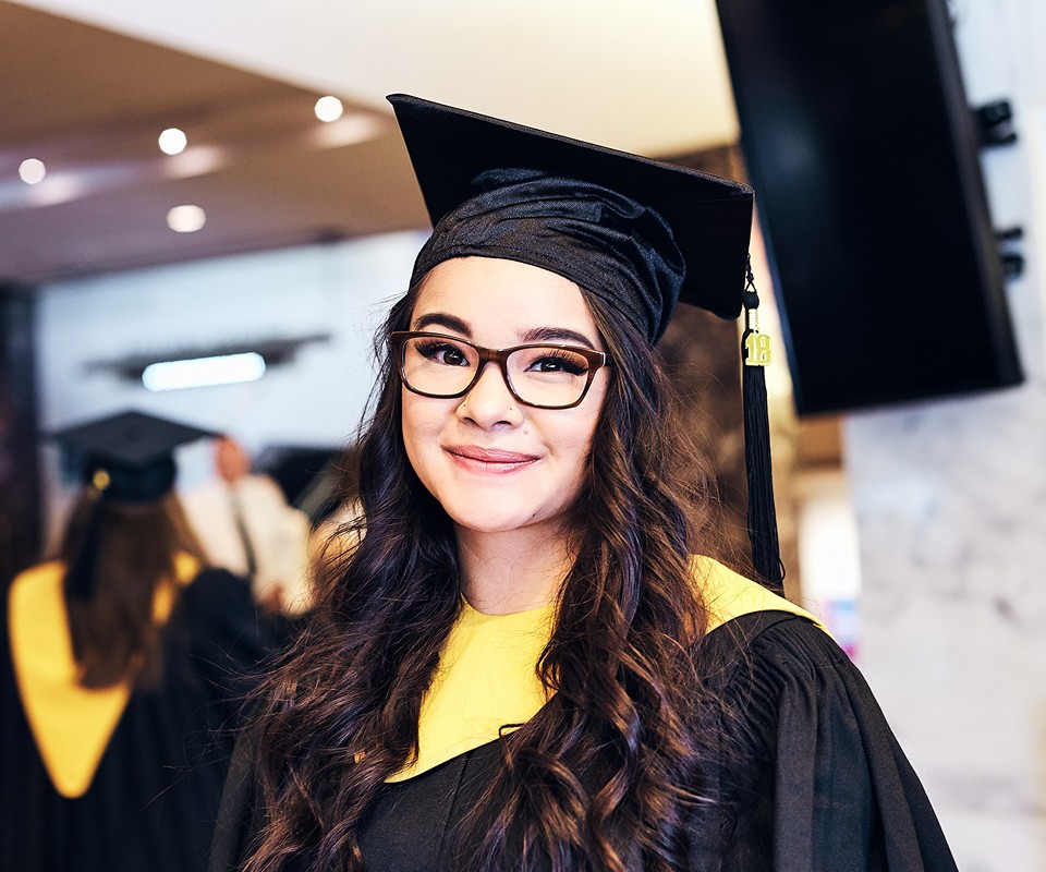 A smiling graduate wearing a black cap and gown with a yellow stole. The background features other graduates in a celebratory setting.