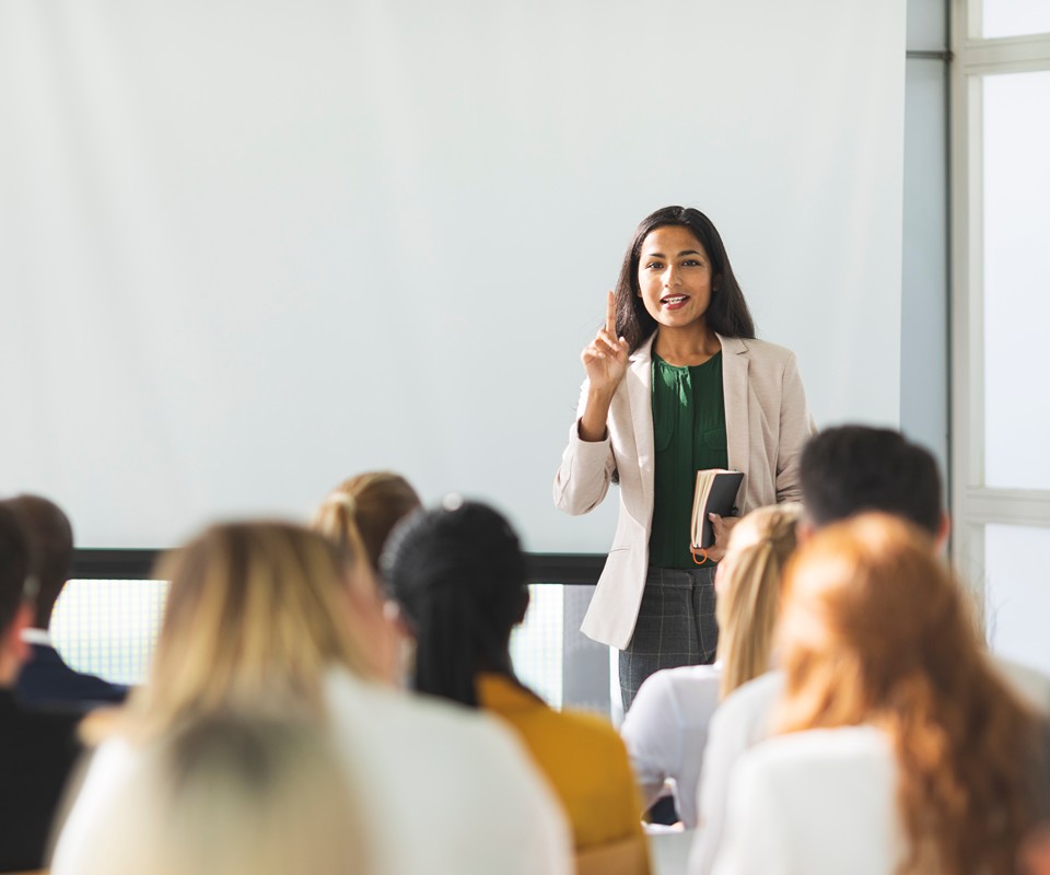 A woman in business attire delivers a presentation to an audience, gesturing with one hand while holding a notebook in the other.