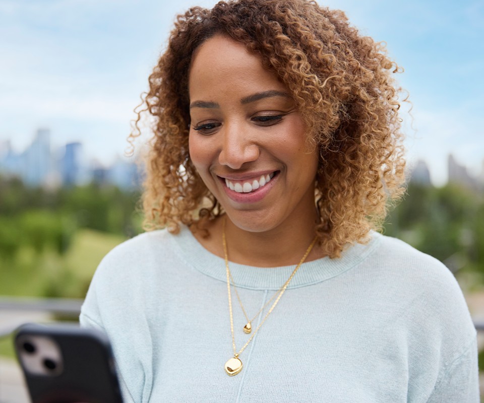 A woman with curly hair smiles while looking at her smartphone outdoors, with a city skyline in the background.