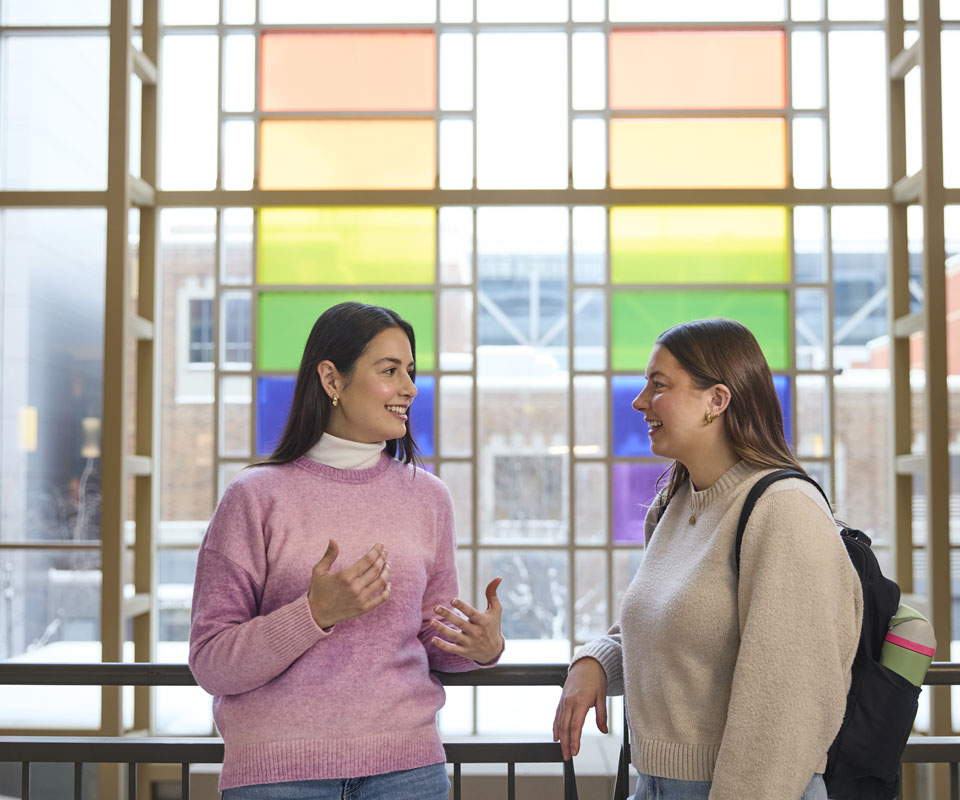Two students engaged in a conversation in front of colorful pride-themed windows inside the Irene Lewis Atrium.