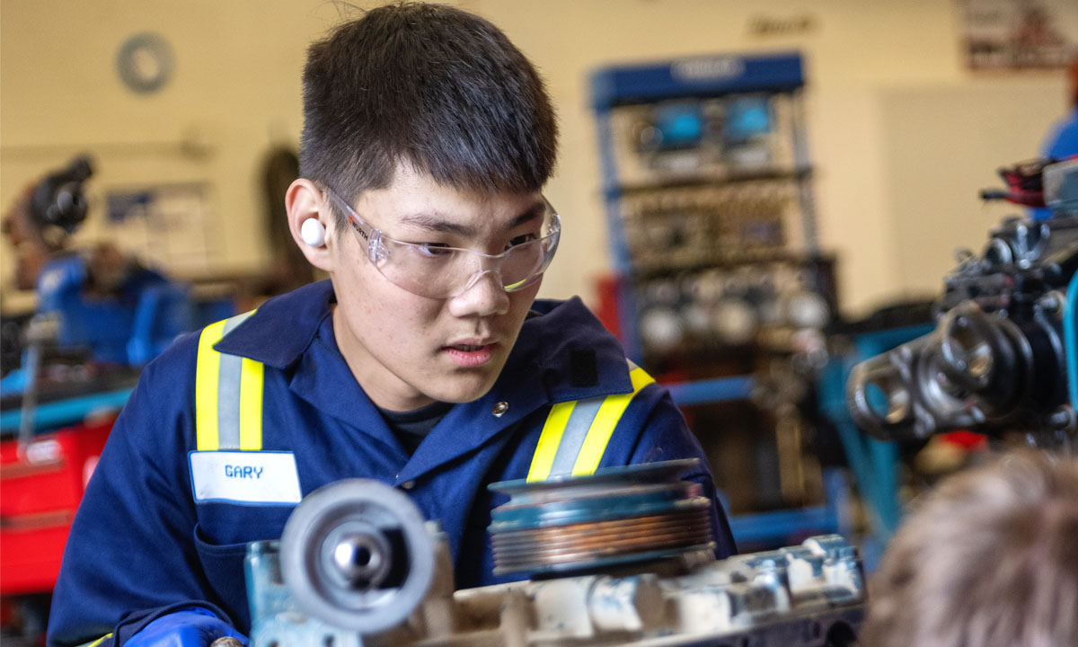 A focused student in a workshop is working on an engine assembly while wearing safety goggles and ear protection.