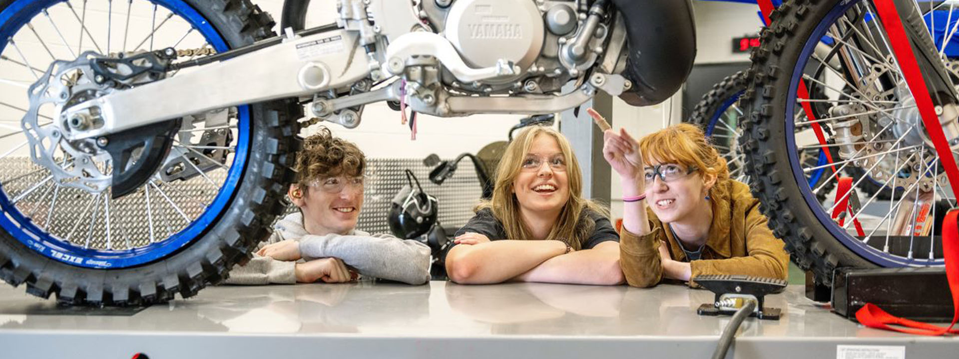 Three students enjoying a hands-on learning experience while working on a motorcycle in a workshop setting.