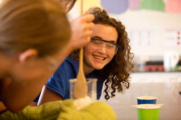 A young SAIT Summer Camper with curly hair smiles. She's wearing safety glasses and watching another camper stir something in a glass beaker.
