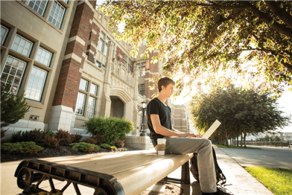A student typing in his laptop sitting on a bench outside