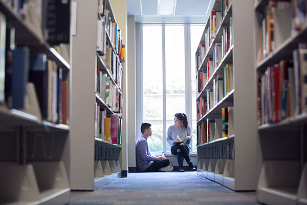 Two students sit on the floor at the end of a bookshelf in the Reg Erhardt Library.