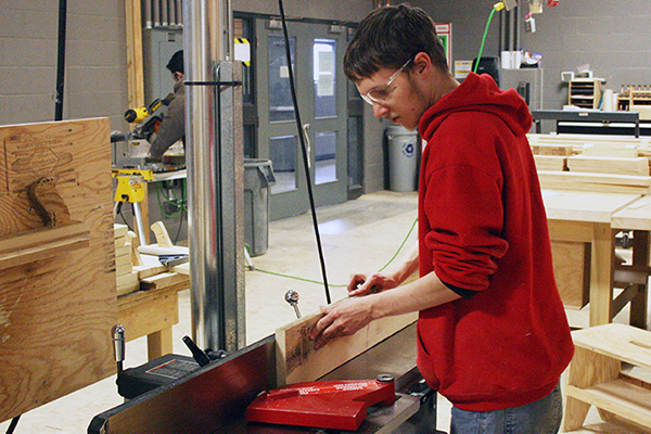 A high school student wearing a red hoodie and safety glasses inspects a plank of wood in a SAIT carpentry lab.