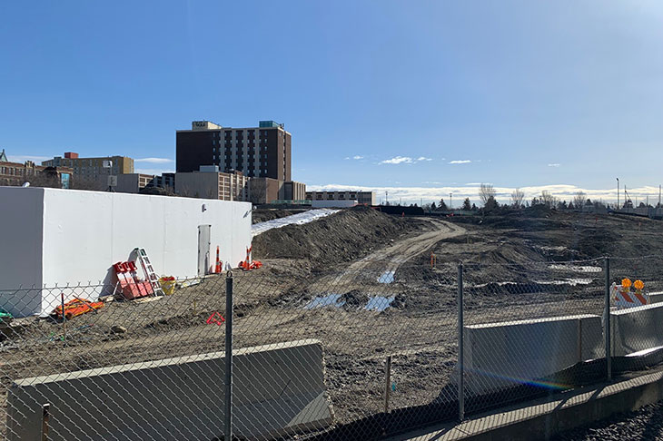 View of Campus Centre construction site from the CTrain platform. Heavy machinery moving earth, with a fence and temporary trailer in the foreground