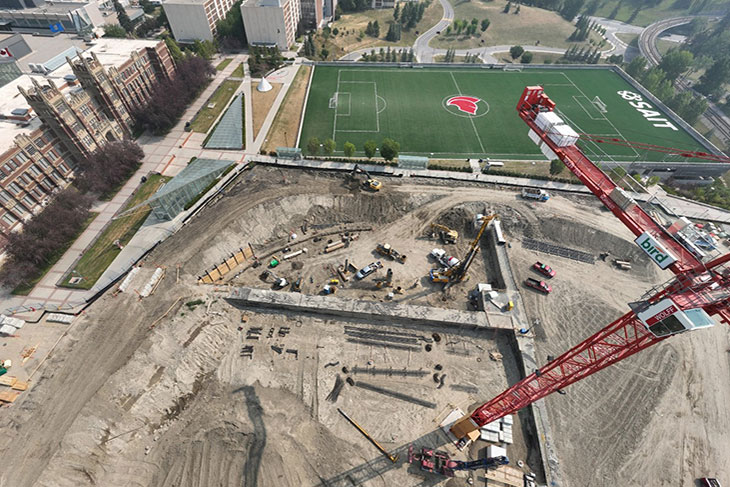 aerial view of the taylor family campus centre construction site on a sunny day, featuring heavy equipment working on dirt mounds, a large crane and portable construction trailers