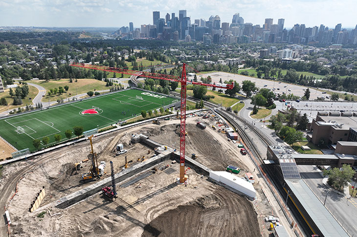 aerial view of the taylor family campus centre construction site on a sunny day, featuring heavy equipment working on dirt mounds, a large crane and portable construction trailers, with a view of downtown calgary in the background