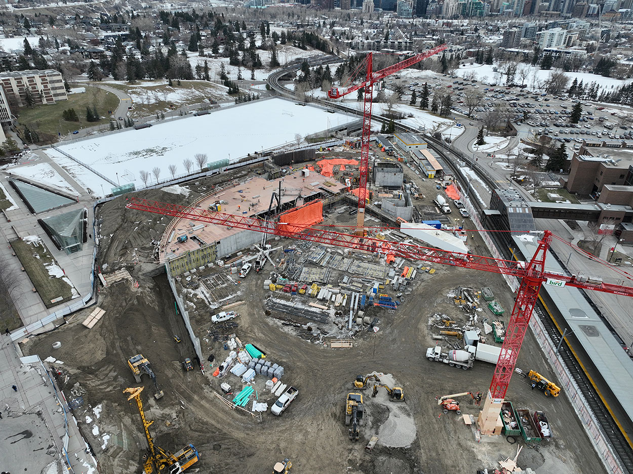 View of Campus Centre construction site. The Cohos Commons field is covered in snow next to the site. Two large red construction cranes and a mix of construction vehicles, equipment and materials can be seen from overhead. The photo also shows the ctrain platform next to the site. and a full parking lot across from the site.