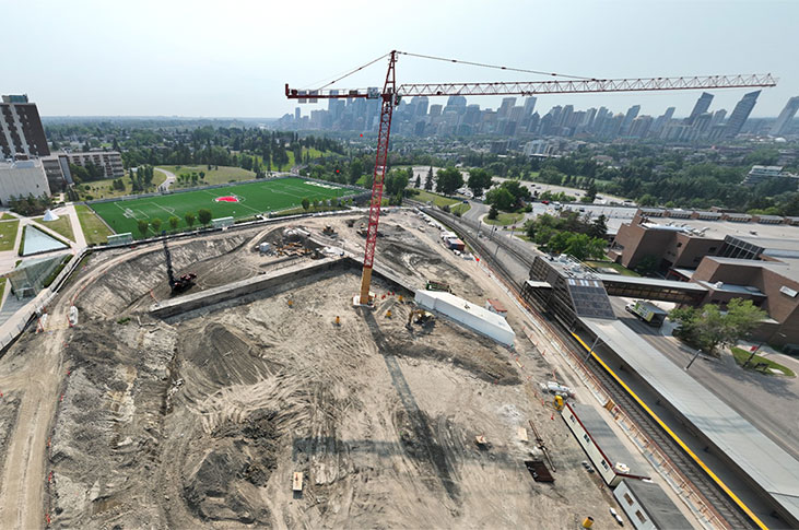 aerial view of the taylor family campus centre construction site on a sunny day, featuring heavy equipment working on dirt mounds, a large crane and portable construction trailers, with a view of downtown calgary in the background
