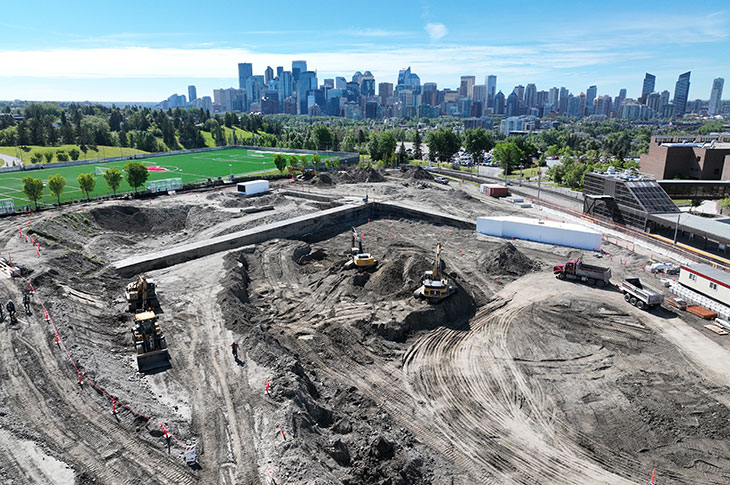 aerial view of the taylor family campus centre construction site on a sunny day, featuring heavy equipment working on dirt mounds and portable construction trailers, with a view of downtown calgary in the background