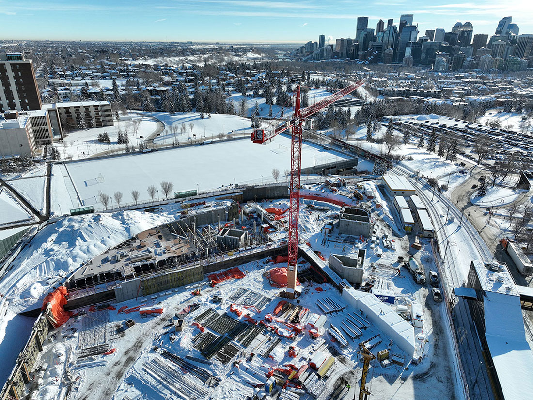 Aerial view of the Taylor Family Campus Centre construction site with snow on the ground, the downtown Calgary cityscape in the background and a large red construction crane.