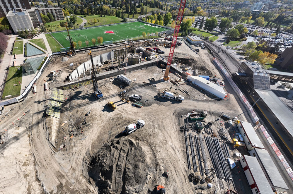 aerial view of the taylor family campus centre construction site on a sunny day featuring heavy equipment working on dirt mounds, a large crane and portable construction trailers