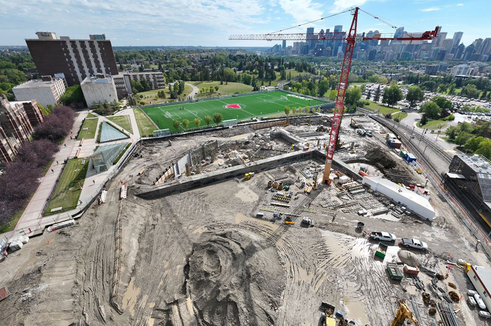 aerial view of the taylor family campus centre construction site on a sunny day with the city of Calgary skyline in the background, featuring heavy equipment working on dirt mounds, a large crane and portable construction trailers