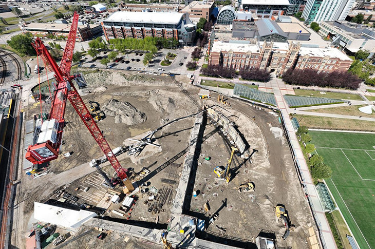 aerial view of the taylor family campus centre construction site on a sunny day, featuring heavy equipment working on dirt mounds, a large crane and portable construction trailers, with a view of Heritage Hall in the background