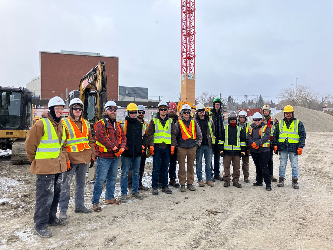 group of SAIT students, employees and contractors wearing safety vests and hard hats on the campus centre construction site with a crane and heavy equipment in the background