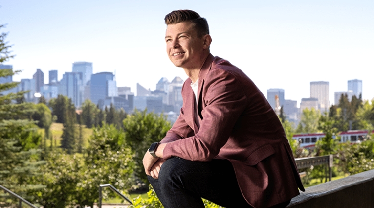 Kyle Bukauskas sitting on concrete wall ledge with city skyline behind him