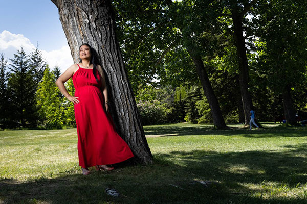 Grace Chantiam wearing a flowing red dress, standing in green trees during the summer.