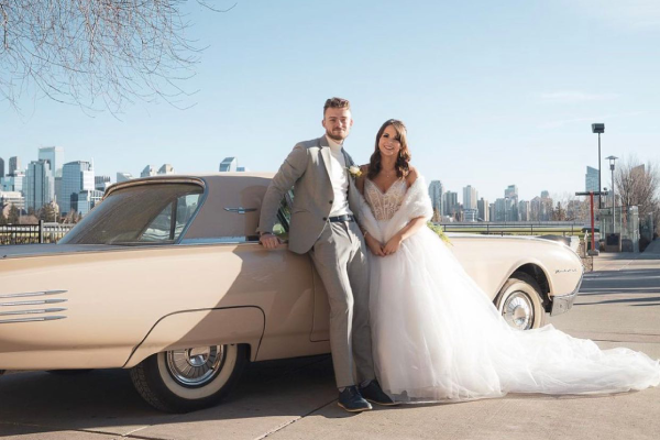 A bride and groom in their wedding attire stand in front of the Calgary skyline from SAIT's campus. They are leaning against a vintage convertible in a pale yellow colour.