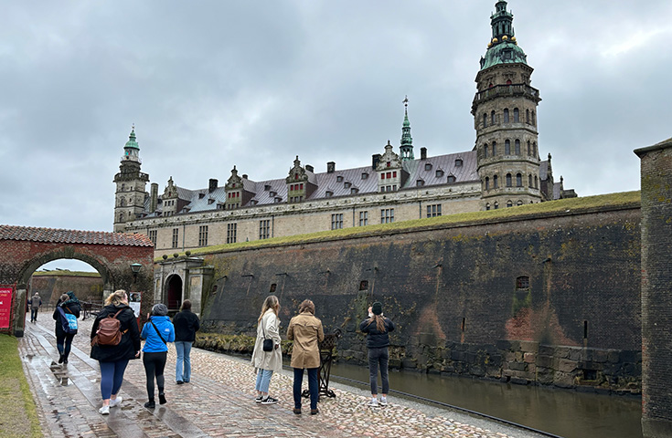 People stand next to a moat on a cobblestone path with a castle in the background.