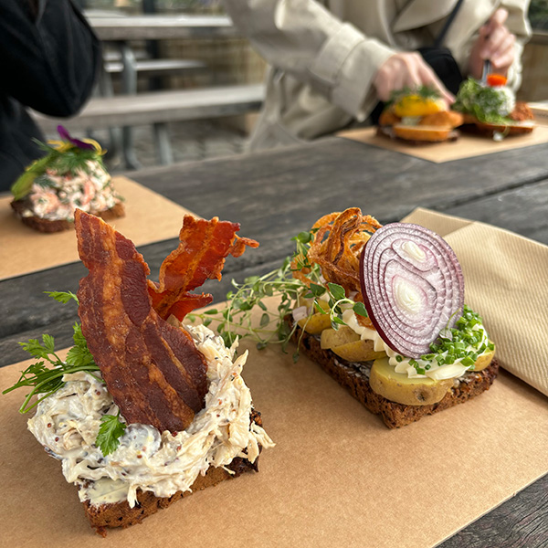 Two open-faced sandwiches sit on a piece of brown paper at a table. The sandwich on the left has two pieces of bacon and the sandwich on the right has a slice of red onion on top.