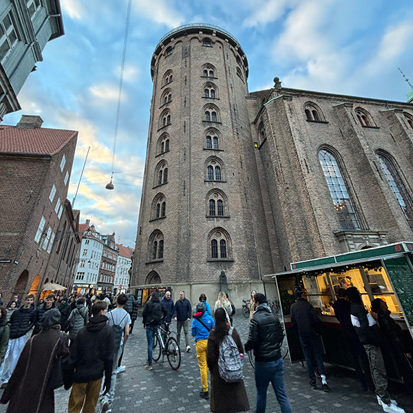 Pedestrians walk through a street with a brown brick tower in the background that’s about eight stories high under a blue sky with some clouds.