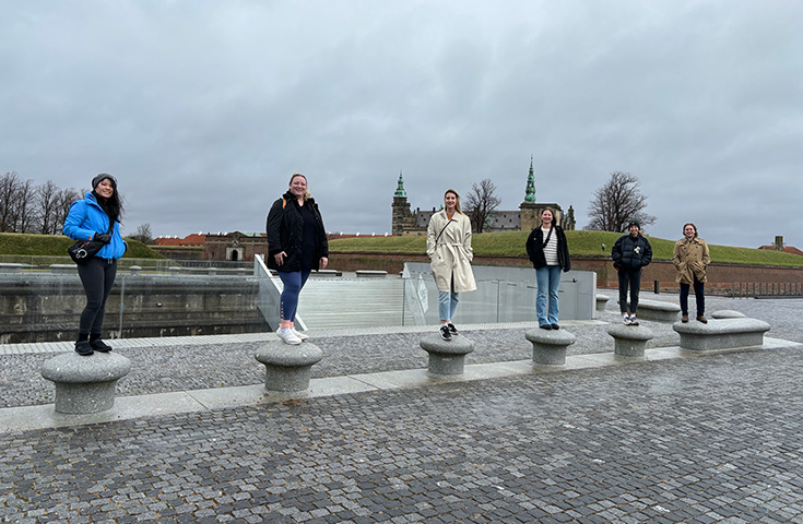 Six people stand in a row on concrete bollards on a cobblestone street with a cloudy sky in the background.