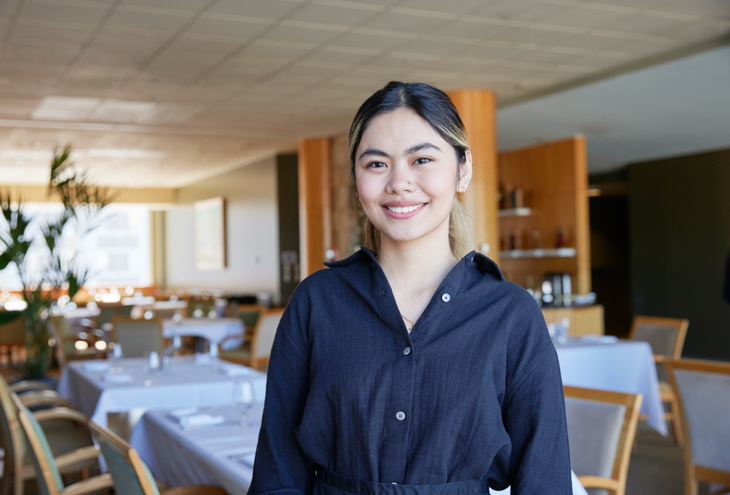 A person smiling at the camera with a restaurant setting in the background