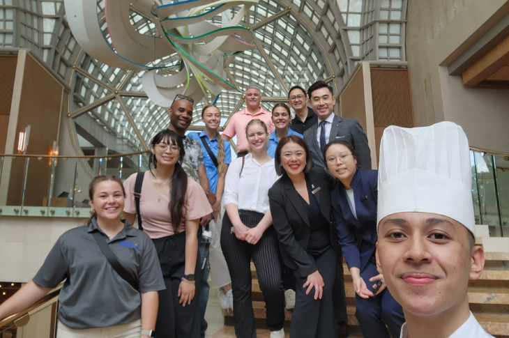 Students stand with staff and a chef from a local hotel in Singapore in front of a large structure inside the hotel lobby.