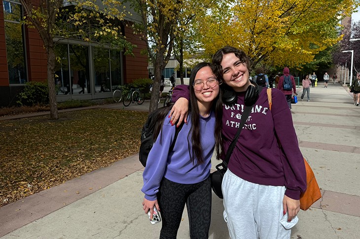 Two people pose for a photo in front of fall foliage, one has her arm around the other's shoulder and both are smiling.