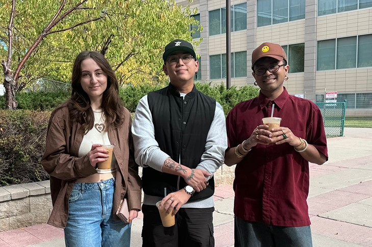 Three people stand outside and pose for a group photo, each holding an iced coffee.