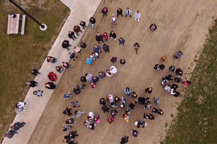 People look up at a camera on a drone over head while standing on a road near a field.