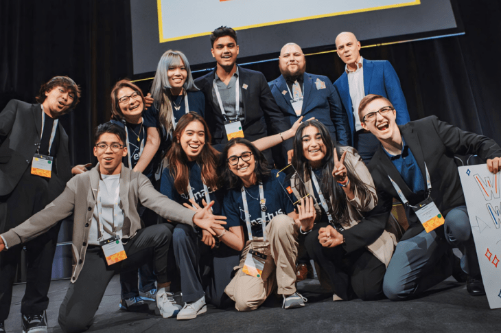 Ten Enactus SAIT students crouch on the ground in front of a black curtain. They are wearing business-casual attire. They are all cheering and smiling at the camera.