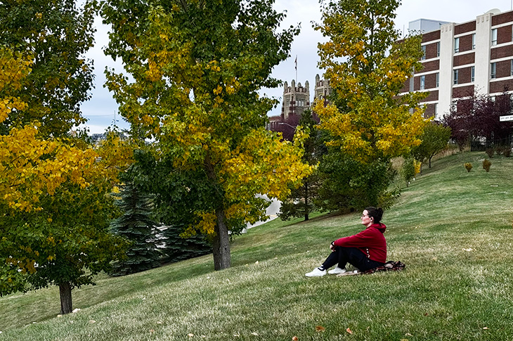 A woman sits on a grassy hill looking into the distance and in the background are yellow trees for fall and Heritage Hall.