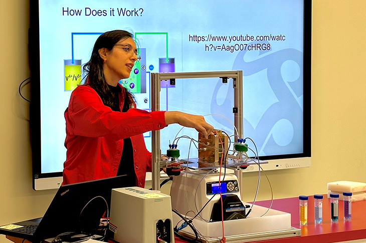 A woman stands at the front of a classroom and a slide that says How does it work? She points to parts of a battery system on a table in front of her.